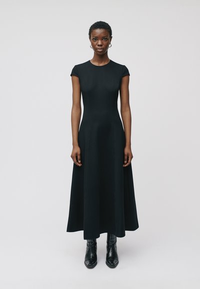 Robe maxi noire ajustée avec des manches courtes à capuchon, un col rond et une silhouette évasée, associée à des bottines noires. Texture lisse.