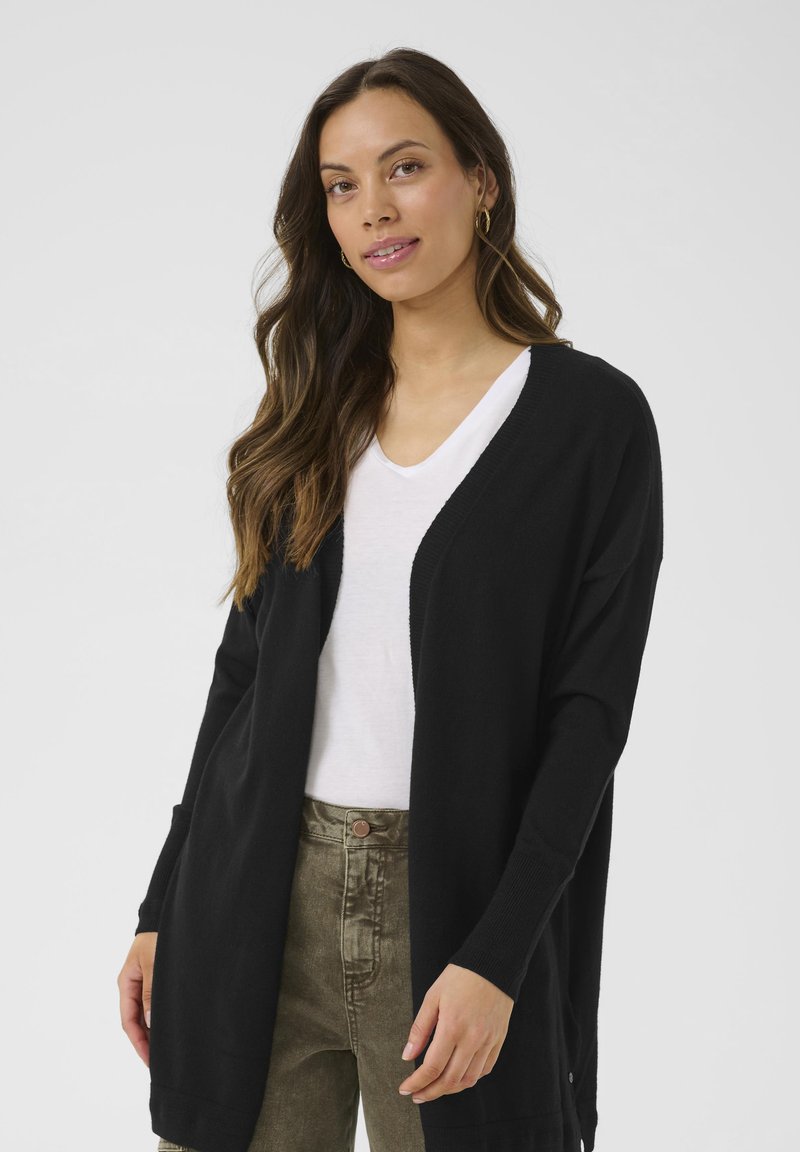 Črn cardigan iz mehkega blaga, odprt spredaj, dolgi rokavi z rebrastimi manšetami, nošen preko bele majice z V-izrezom, dopolnjen z olivno zelenimi hlačami.