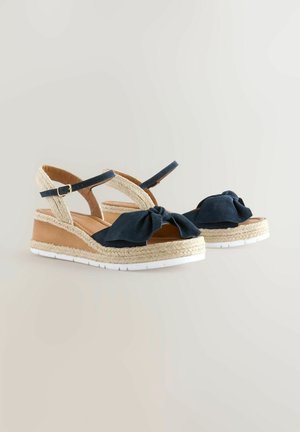 Tumesinine wedgesandaalid, millel on sõlmitud lipsu detail, kudumisest valmistatud jute aktsendid ja valged kummist tallad. Reguleeritavad pahkluu rihmad mugavuse tagamiseks.