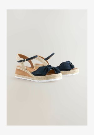 Tumesinine wedgesandaalid, millel on sõlmitud lipsu detail, kudumisest valmistatud jute aktsendid ja valged kummist tallad. Reguleeritavad pahkluu rihmad mugavuse tagamiseks.