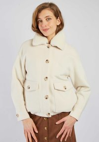 Biała kurtka z mieszanki wełny z dużymi guzikami, luźnym krojem, dwoma kieszeniami z przodu i kołnierzem z futerka. Noszona na brązowej spódnicy.