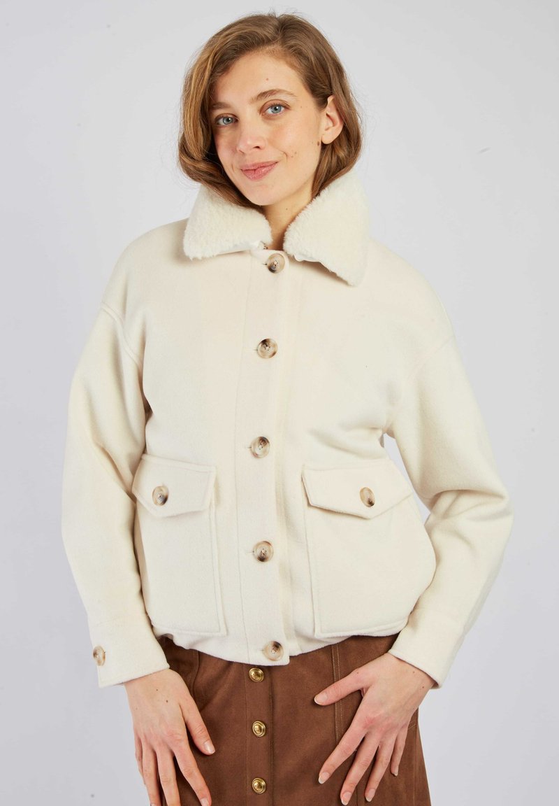 Biała kurtka z mieszanki wełny z dużymi guzikami, luźnym krojem, dwoma kieszeniami z przodu i kołnierzem z futerka. Noszona na brązowej spódnicy.