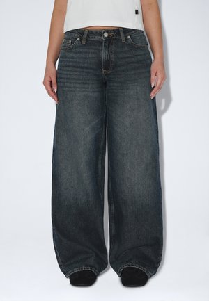 Osoba nosząca ciemne, szerokie spodnie jeans z wysokim stanem i czarne buty, stojąca na jednolitym jasnym tle.
