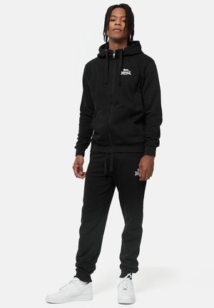 Sort zip-up hoodie med snore og logo på brystet, parret med matchende sorte joggers. Bæres med hvide sneakers.