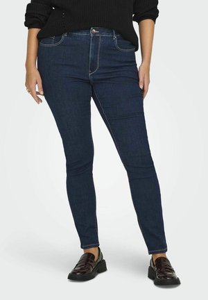 Jean skinny bleu foncé avec une texture en denim lisse, doté d'une fermeture à bouton à l'avant et d'accents de couture. Porté avec des mocassins noirs.