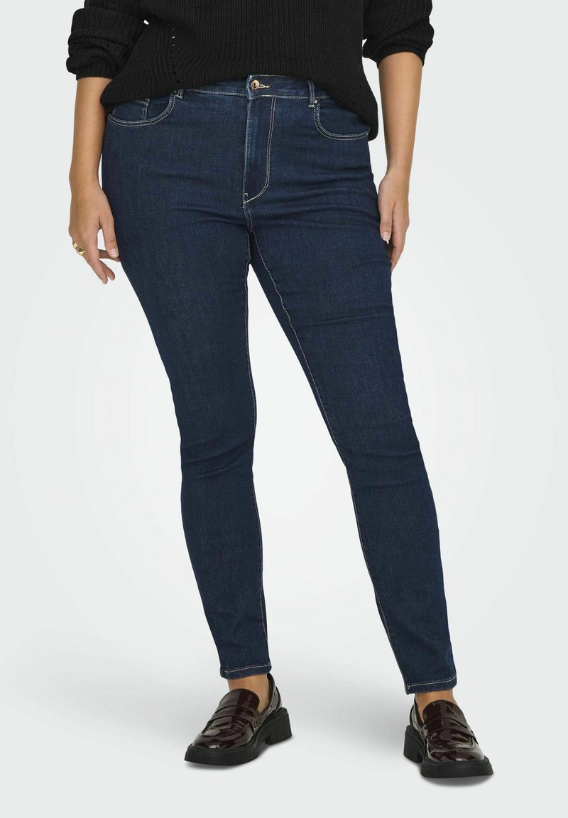 Jeans skinny de culoare albastru închis, cu o textură netedă de denim, dotate cu un sistem de închidere cu nasture în față și accente de cusătură. Purtați cu loafers negri.