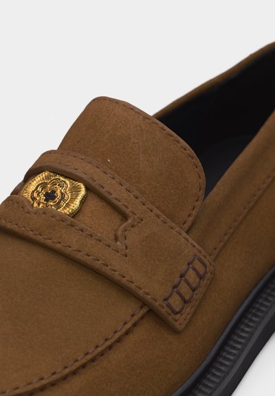 Mocassin en suède marron avec une semelle noire et un détail décoratif en métal doré sur la bride supérieure.