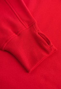 Punane dressipluus, millel on sooniline mansett ja pöidlaharu detail, valmistatud pehmest kangast sileda tekstuuriga. Suumi vaade manseti piirkonnale.