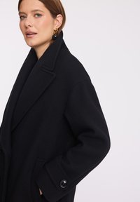 Czarny wełniany płaszcz z wyciętym kołnierzem, o oversize'owym kroju, z bocznymi kieszeniami i guzikami na rękawach. Gładka tekstura i minimalistyczny design.