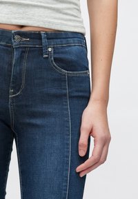Temno modre denim jeans z ozkim krojem. Ponašajo se z zadrgo, gumbnim zapiranjem, zankami za pas in belim kontrastnim šivom ob šivih.