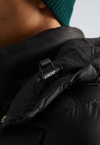 Schwarze Daunenjacke mit glatter Textur, ausgestattet mit einem grafischen Logo-Zipper und kontrastierenden Nähten, teilweise am Kragen geöffnet.