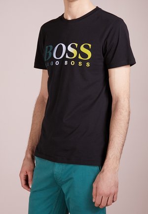 Mężczyzna w czarnej koszulce Hugo Boss z wielokolorowym logo i turkusowych spodniach, stojący na beżowym tle.
