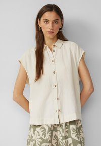 Cremefarbendes, kurzärmeliges Blusenshirt mit Knopfleiste und subtiler Textur, kombiniert mit gemusterten grünen Shorts mit floraler Gestaltung.
