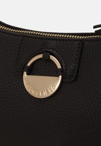 Czarna torba z teksturowanej skóry z ozdobnym, okrągłym elementem w kolorze złotym z wytłoczonym logo marki oraz zamkiem błyskawicznym u góry.