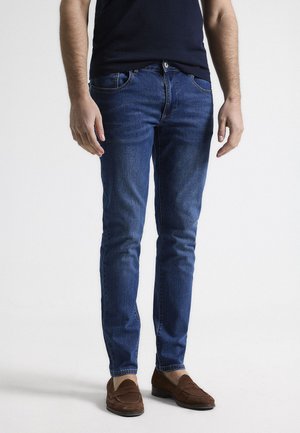 Jeans skinny azul oscuro con cinco bolsillos, cintura de altura media, sutiles detalles de costura y una textura de denim suave. Combinados con zapatos marrones de tipo slip-on.