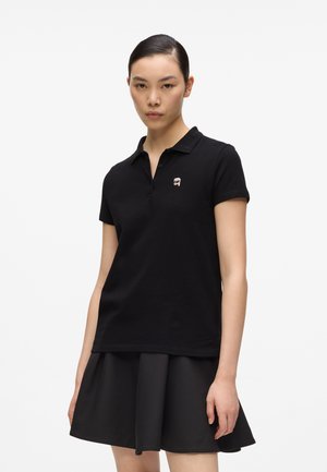 Jeune femme portant un polo noir à manches courtes avec un petit logo brodé et une jupe noire, debout devant un fond clair.