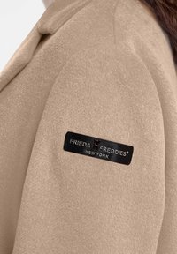 Beige, zacht-textuur jas met een schoudernaad; voorzien van een zwart stoffen label met de tekst "FRIEDA® FREDDIES NEW YORK."