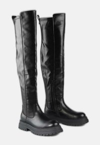 Botas de cuero negras por encima de la rodilla con suelas gruesas y estriadas y cremalleras laterales, de pie en posición vertical sobre un fondo blanco.