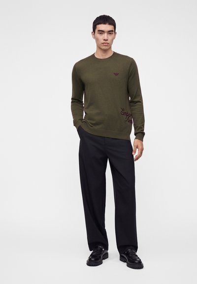 Jeune homme portant un pull vert olive avec un pantalon noir et des chaussures noires, debout avec une main dans la poche devant un fond uni.