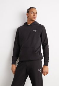 Felpa nera con cappuccio realizzata in tessuto morbido, dotata di tasca a marsupio e di un logo bianco Puma sul petto. Accompagnata da pantaloni neri coordinati.