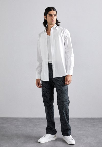 White button-up shirt with a black logo on the collar, paired with black pants featuring a repeated logo pattern and white sneakers.