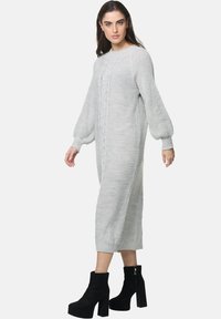 Un lungo abito a maglia grigio chiaro con dettagli a treccia, maniche a palloncino e una vestibilità rilassata, abbinato a stivali neri con tacco alto alla caviglia.