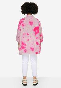 Kimono lungo e oversize in un motivo tie-dye rosa su sfondo chiaro, con maniche larghe e una vestibilità rilassata. Indossato sopra pantaloni bianchi.