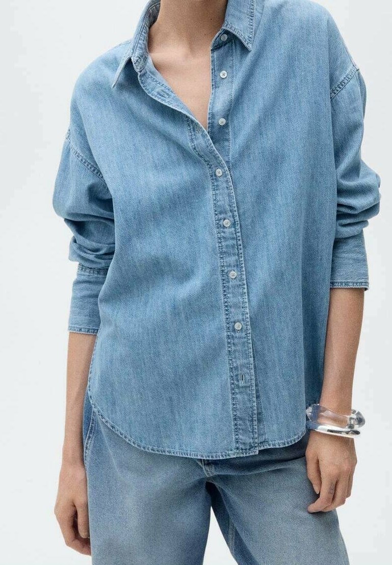 Femme portant une chemise en denim bleu clair à boutons avec manches retroussées et un jean en denim assorti, agrémentée de bracelets transparents et argentés.
