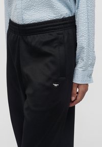 Άτομο που φοράει μαύρο παντελόνι Carhartt WIP με τσεπάκι με φερμουάρ και γαλάζιο υφασμάτινο πουκάμισο με μακριά μανίκια, το χέρι ακουμπάει στο πλευρό.