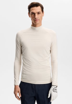 Mężczyzna w beżowej bluzce z długim rękawem i stójką, ciemnych spodniach oraz białej golfowej rękawiczce na lewej ręce, stojący na jednolitym tle.