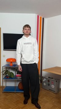 Jonge man staat in een kamer, draagt een witte hoodie en zwarte broek, met een basketbal, een oranje lamp, een tv en een koffer in de buurt.