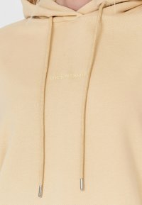 Béžová mikina s kapucí s texturovaným povrchem a kovovými detaily na stahovacích šňůrkách. Na přední straně je vyšitý nápis "COTTON CANDY" světlejší barvou.