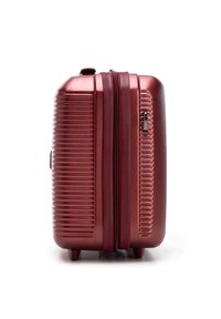 Červený kovový kufr s tvrdou skořápkou postavený ve vzpřímené poloze, s vodorovnými drážkami, zipem a dvěma malými kolečky na bílém pozadí.