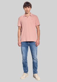 Ljusrosa polo skjorta med krage, parat med blå denimjeans och beige sneakers. Skjortan har ett litet emblem på bröstet.