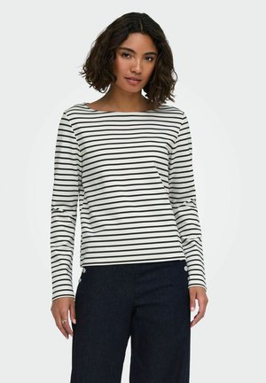 Vrouw draagt een wit shirt met lange mouwen met zwarte horizontale strepen en een donkere, hoog geplaatste broek met knopen op de heupen.