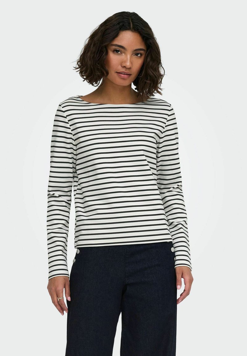 Femme portant un haut blanc à manches longues avec des rayures horizontales noires et un pantalon sombre taille haute avec des boutons sur les hanches.