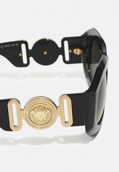 Svarta solglasögon med kraftiga bågar, som har en gyllene cirkulär detalj med ett dekorativt mönster och förlängda gyllene accenter på bågarna.