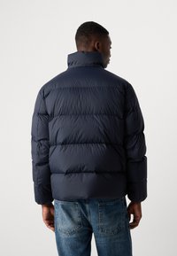 Navy puffer jacket featuring a high collar, quilted design, and soft, smooth texture. Paired with light blue denim jeans.