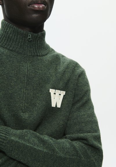 Zelený pletený svetr s vysokým límcem a půlzipem, na hrudi s bílou vyšívanou logem "W". Měkká textura a žebrované manžety.
