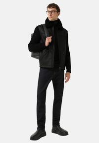 Zwarte leren vest met een nep bontkraag, gecombineerd met een zwarte coltrui, donkere spijkerbroek en zwarte enkellaarsjes. Accessoire: grijze rugzak.