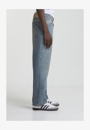 Oseba, oblečena v svetlo modre jeans hlače s pravimi hlačnicami in bele Adidas superge s črnimi črtami, stoji bočno na sivem tlaku.