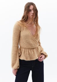 Brun texturerad omslagströja med långa, voluminösa ärmar och peplummidja, som bärs tillsammans med mörkblå denimjeans.