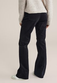 Persona che indossa pantaloni in velluto a coste neri, un maglione beige chiaro e scarpe slip-on bianche, in piedi su uno sfondo neutro.