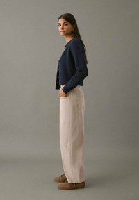 Cardigan de punto azul marino con botones combinado con pantalones de denim crema, que cuentan con bolsillos laterales. El modelo lleva zapatos casuales marrones y el cabello suelto.
