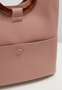Sac à main en cuir rose blush avec des poignées rondes en écaille de tortue et une poche avant fermée par un bouton pression.