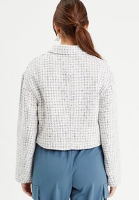 Kurz geschnittene Tweedjacke in Weiß mit blauen und marineblauen Fäden, mit einem Reverskragen und langen Ärmeln. Die Rückansicht zeigt die Textur und Passform.