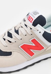 Beige und marineblaue New Balance 574 Sneakers mit einem roten Logo, aus Mesh und Wildleder, ausgestattet mit weißen Akzenten und kontrastierenden Schnürsenkeln.
