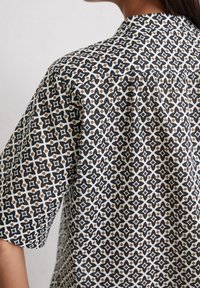 Chemise à manches courtes avec un motif géométrique en noir, blanc et marron, dotée d'un col classique et de détails plissés au dos.