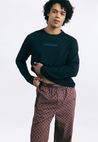 Melna garām piedurknēm krekls ar "Calvin Klein" logo; sarkanas rakstotas pidžamu bikses ar elastīgu jostu un logo detaļu. Kūrīgs mati.