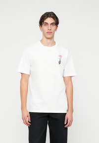 T-shirt en coton blanc à manches courtes. Présente un design de rose rose et un accent noir et blanc sur le côté gauche de la poitrine. Coupe simple et décontractée.